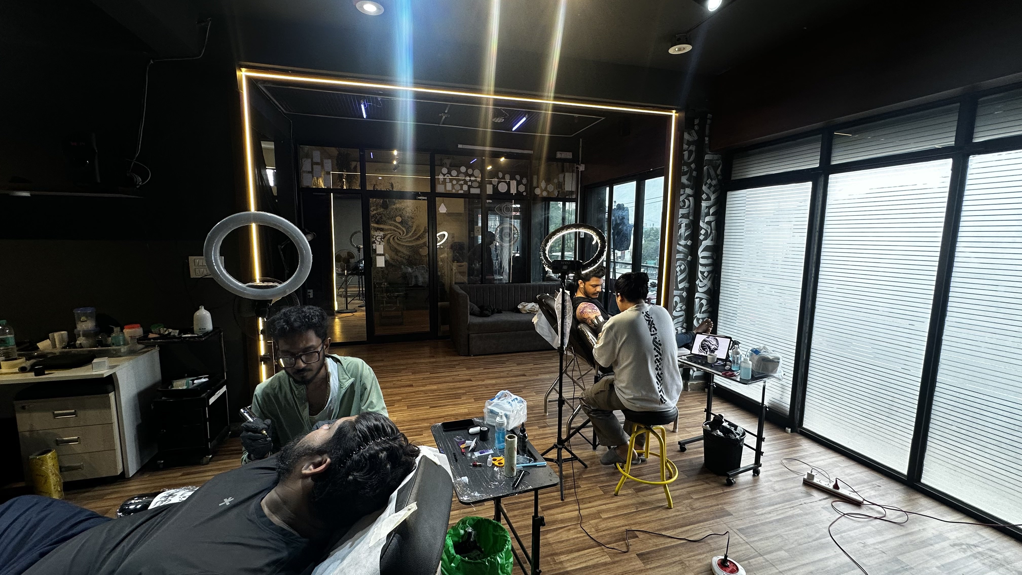 Both artists tattooing clients simultaneously — wide studio view with golden LED frame
